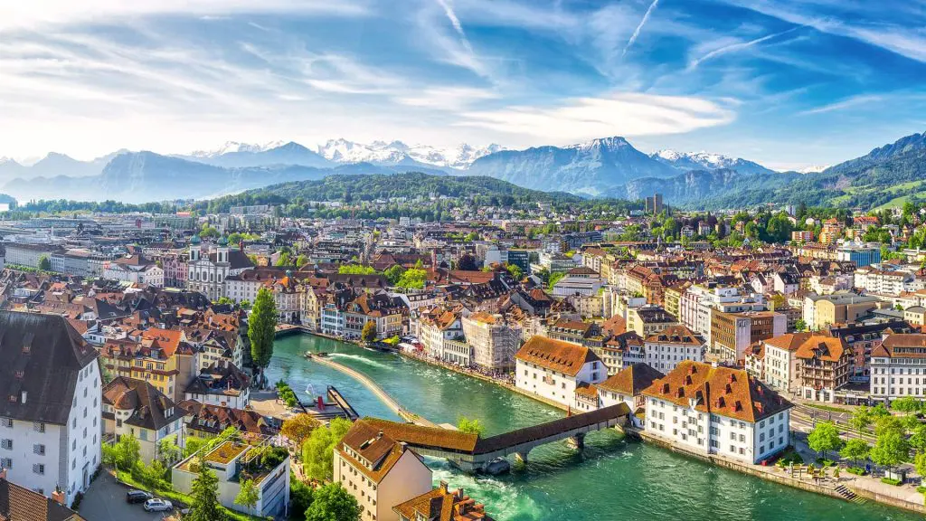 Panoramic view of a swiss city with historic architecture along a river and the alps in the background.