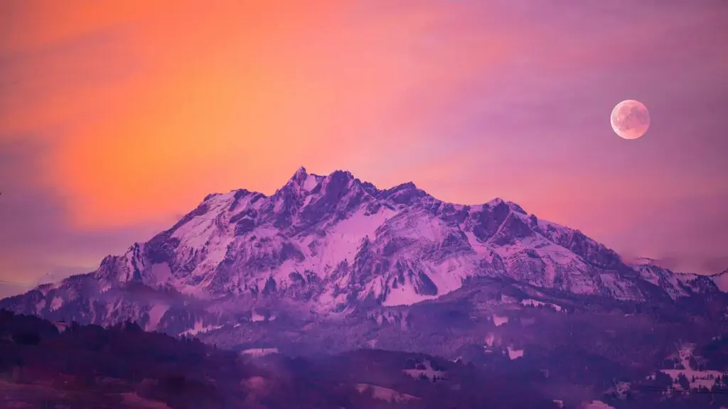 Snow-covered mountain peaks under a vibrant orange and purple sky with a full moon.