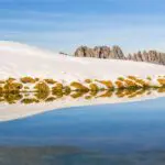 Snowy slopes and grassy patches reflected in a calm lake, with jagged mountain peaks in the background under a clear blue sky—a perfect scene to include on your Switzerland itinerary.