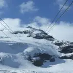 Snow-covered mountain peak with cables stretching across the sky. Cloudy blue sky in the background.