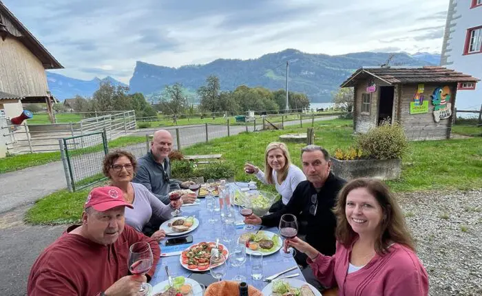 Six people sitting at an outdoor table enjoying a meal with wine. Scenic background with mountains and buildings.