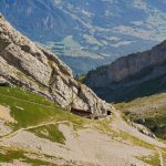 A red cogwheel train travels along a mountain track through rocky terrain, with a valley and village visible in the background—an iconic scene for travellers who may also wonder about customs like tipping in Switzerland.